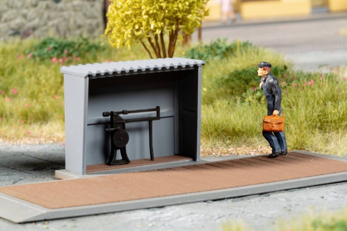 Un motif de train miniature représente un employé de chemin de fer avec un sac devant un petit abri doté d'un mécanisme rotatif, entouré de végétation verdoyante.