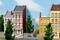 Trois maisons mitoyennes colorées avec des commerces au rez-de-chaussée, bordées d'arbres, sur un fond de ciel bleu et de nuages.