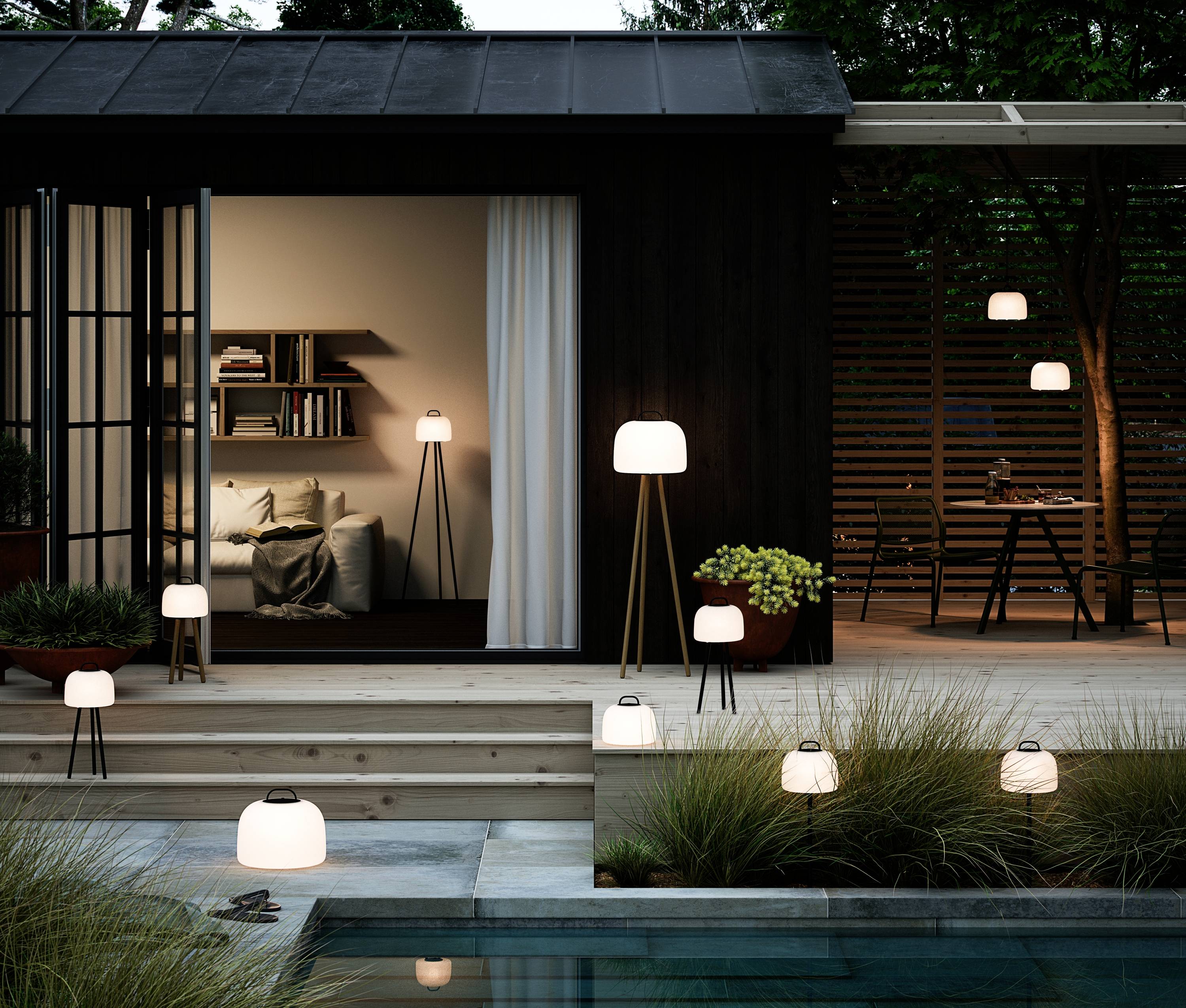 Maison moderne illuminée au crépuscule avec plusieurs lampes sur la terrasse et dans le jardin, vue sur le salon avec canapé et étagère.