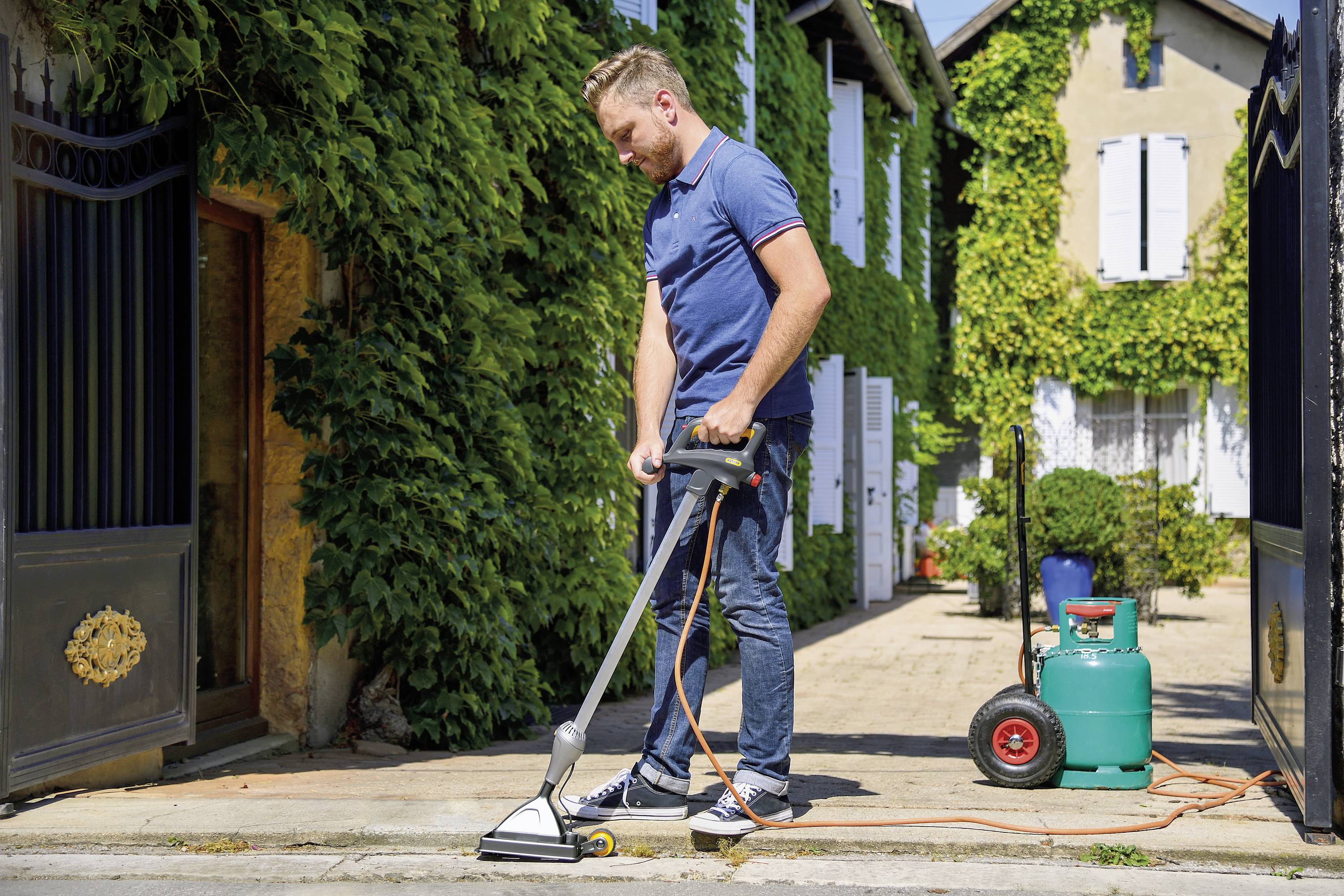 Un homme nettoie le pavage devant une maison entourée de plantes à l'aide d'un nettoyeur haute pression. Des bouteilles de gaz vertes sont visibles en arrière-plan.