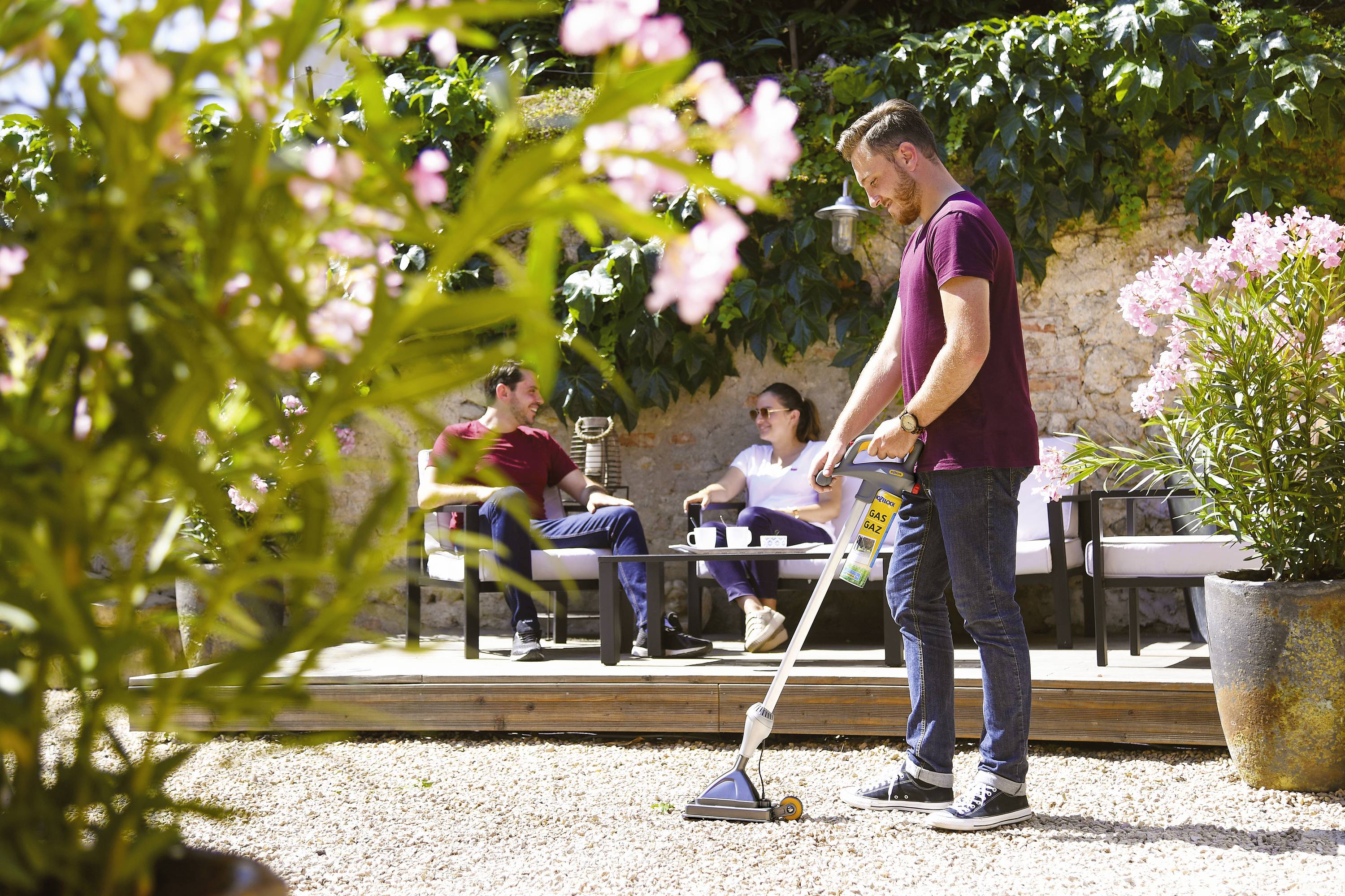 Un homme nettoie le sol en gravier dans un jardin à l'aide d'un appareil. À l'arrière-plan, deux personnes sont assises sur un canapé sous un mur couvert de plantes.