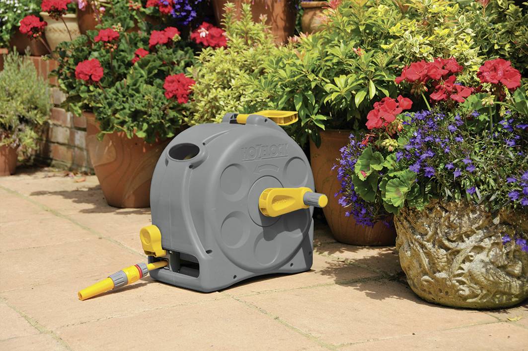 Une boîte de rangement grise pour tuyau de jardin avec des poignées jaunes est posée sur une terrasse, entourée de plantes en fleurs dans différents pots.