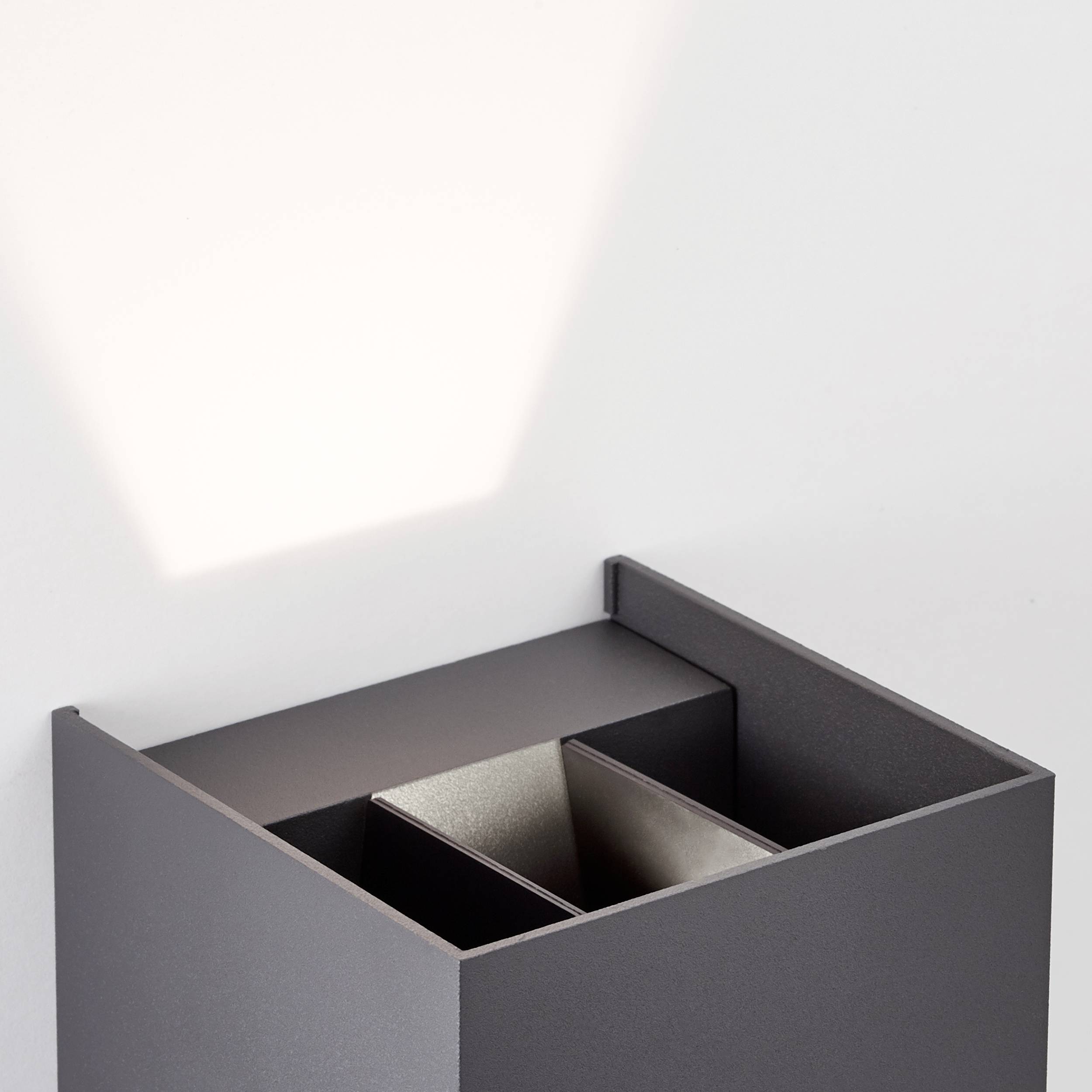 Une petite boîte métallique carrée en matériau gris foncé avec des compartiments ouverts, éclairée par un rayon de lumière provenant d'en haut à gauche.
