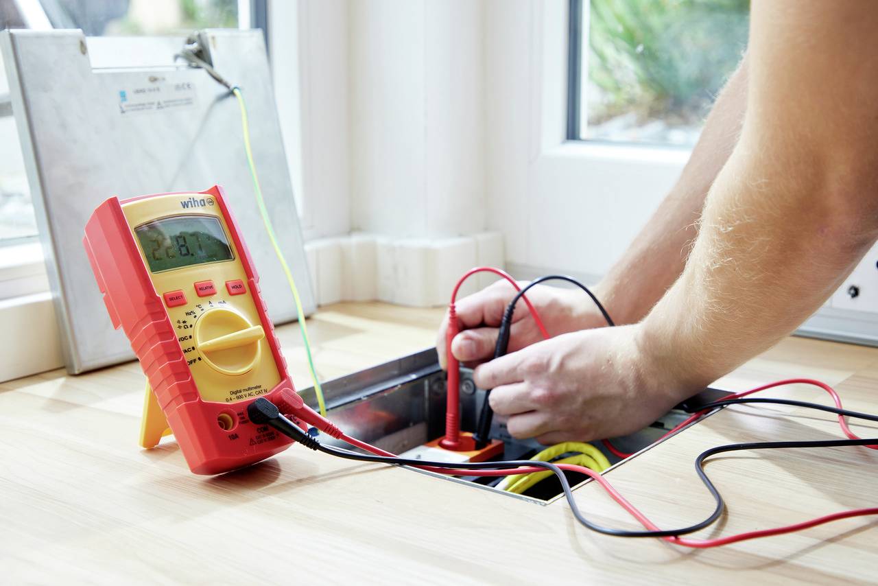 Une personne mesure la tension électrique dans un puits de visite à l'aide d'un multimètre numérique. Les câbles sont branchés.