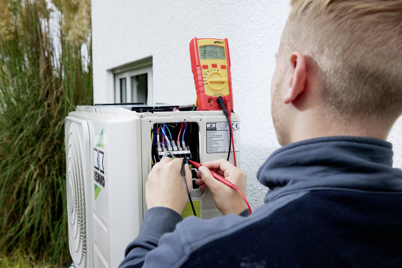 Un technicien vérifie les connexions de câbles d'une pompe à chaleur installée sur un mur extérieur à l'aide d'un multimètre.