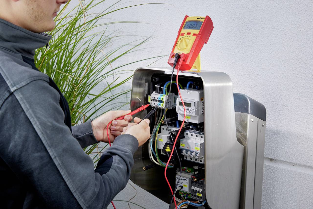 Une personne vérifie les connexions électriques dans un tableau électrique ouvert à l'aide d'un multimètre.
