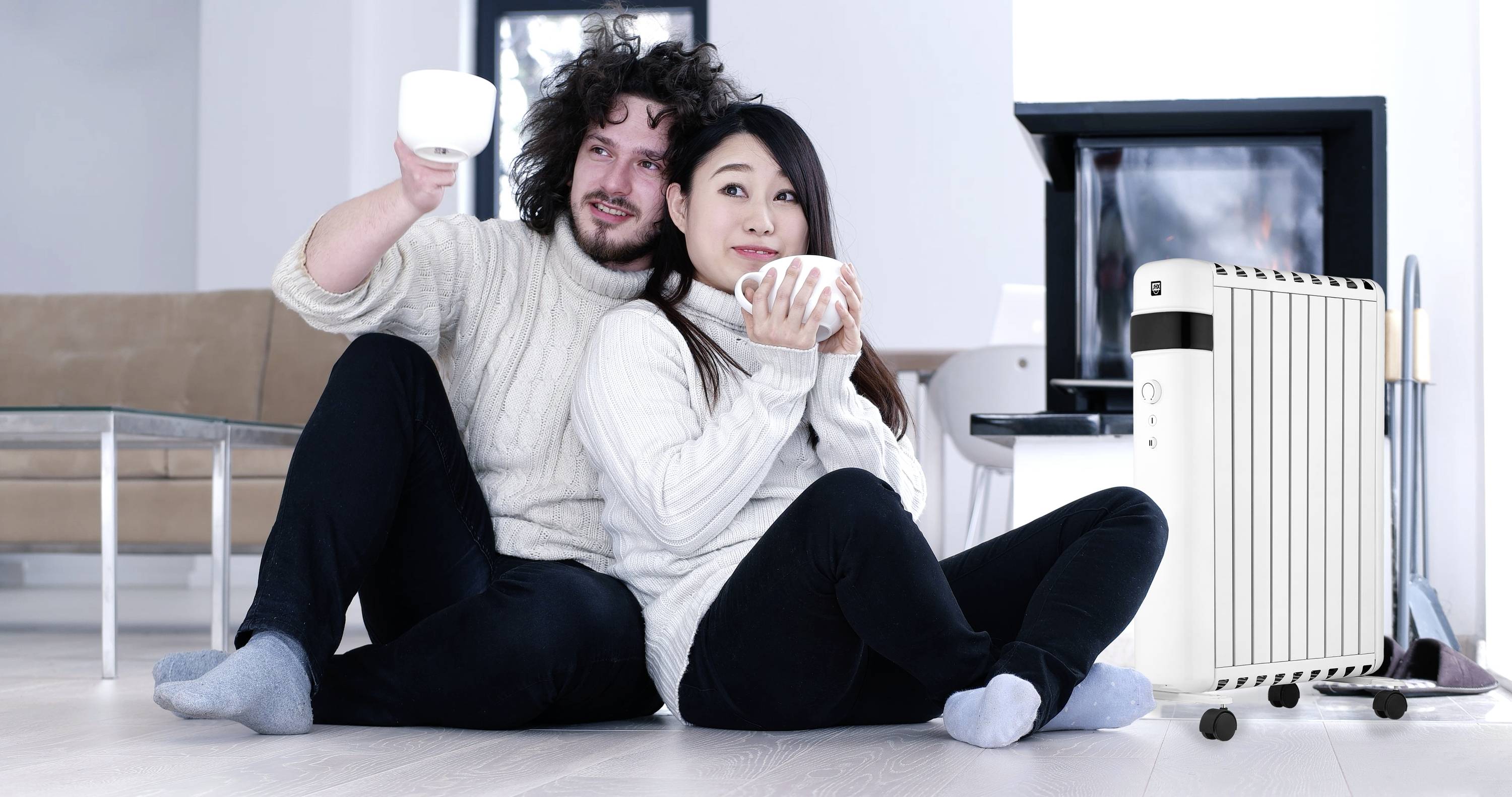 Un couple est assis sur le sol du salon, souriant et tenant des tasses. Un radiateur est à côté d'eux. La scène semble chaleureuse et confortable.