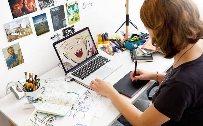 Une personne dessine sur une tablette graphique à un bureau, entourée de croquis et de matériel artistique. Un écran d'ordinateur portable affiche une illustration colorée.