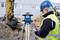 Un ouvrier portant un casque de chantier et un gilet de sécurité utilise un instrument de mesure sur un chantier, avec un pelleteuse en arrière-plan.