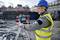 Un ouvrier portant un casque bleu et un gilet de sécurité jaune manipule un appareil de mesure sur un chantier avec des travaux de terrassement en arrière-plan.