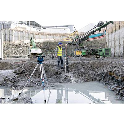 Un ouvrier du bâtiment en tenue de protection utilise un équipement lourd sur un grand chantier, avec une foreuse et un instrument de mesure au premier plan.