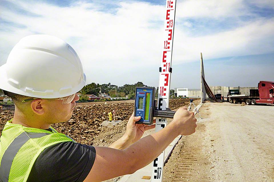 Un ouvrier portant un casque de chantier et un gilet de sécurité mesure le profil du terrain sur un chantier à l'aide d'un niveau et d'une mire.