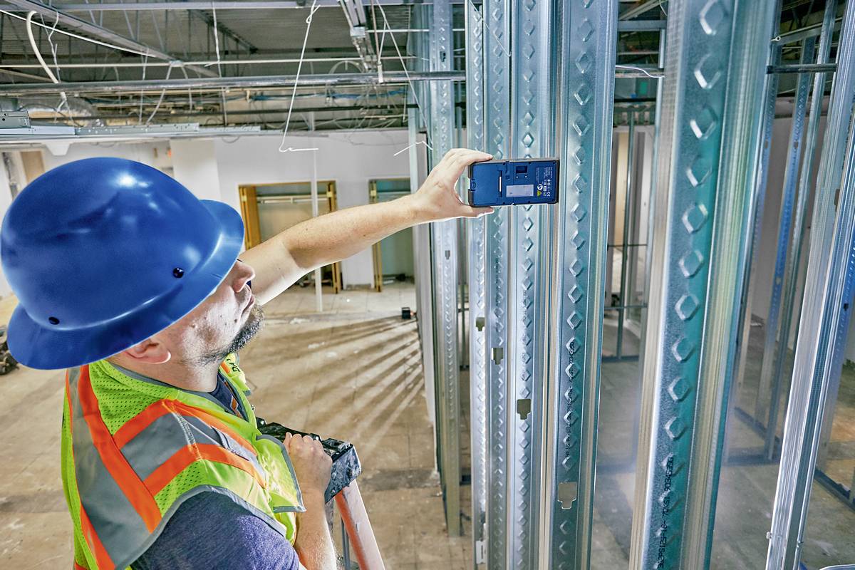 Un ouvrier portant un casque bleu et un gilet de sécurité mesure des poutres métalliques sur un chantier à l'aide d'un télémètre laser.