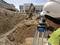 Un ouvrier mesure un chantier à l'aide d'un instrument de topographie, tandis qu'un pelleteuse déplace de la terre en arrière-plan.