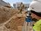 'Un ingénieur en génie civil portant un casque de chantier utilise un appareil de mesure sur un chantier, tandis qu'un excavateur travaille en arrière-plan.'