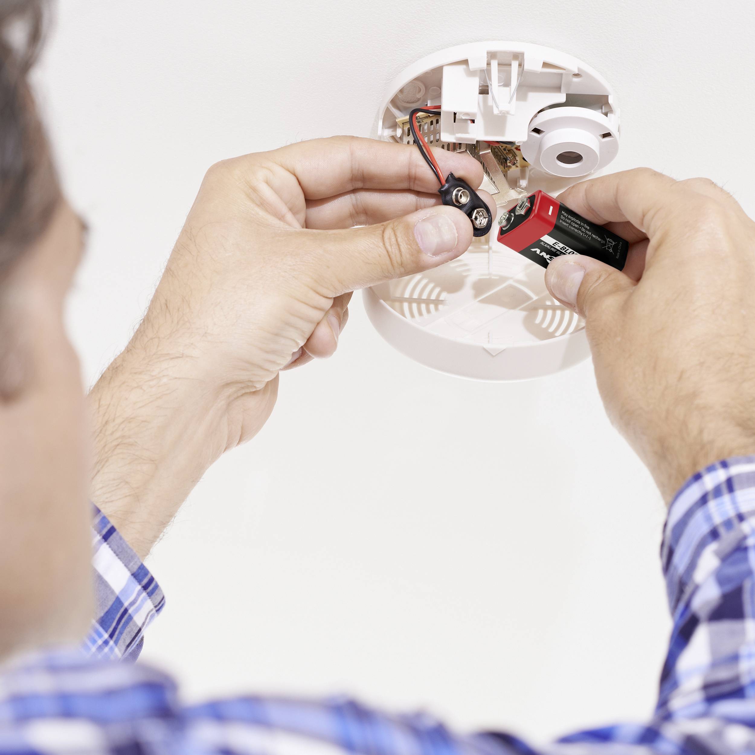 Une personne installe une pile dans un détecteur de fumée au plafond. Les mains sont visibles, les détails du détecteur de fumée sont mis au point.