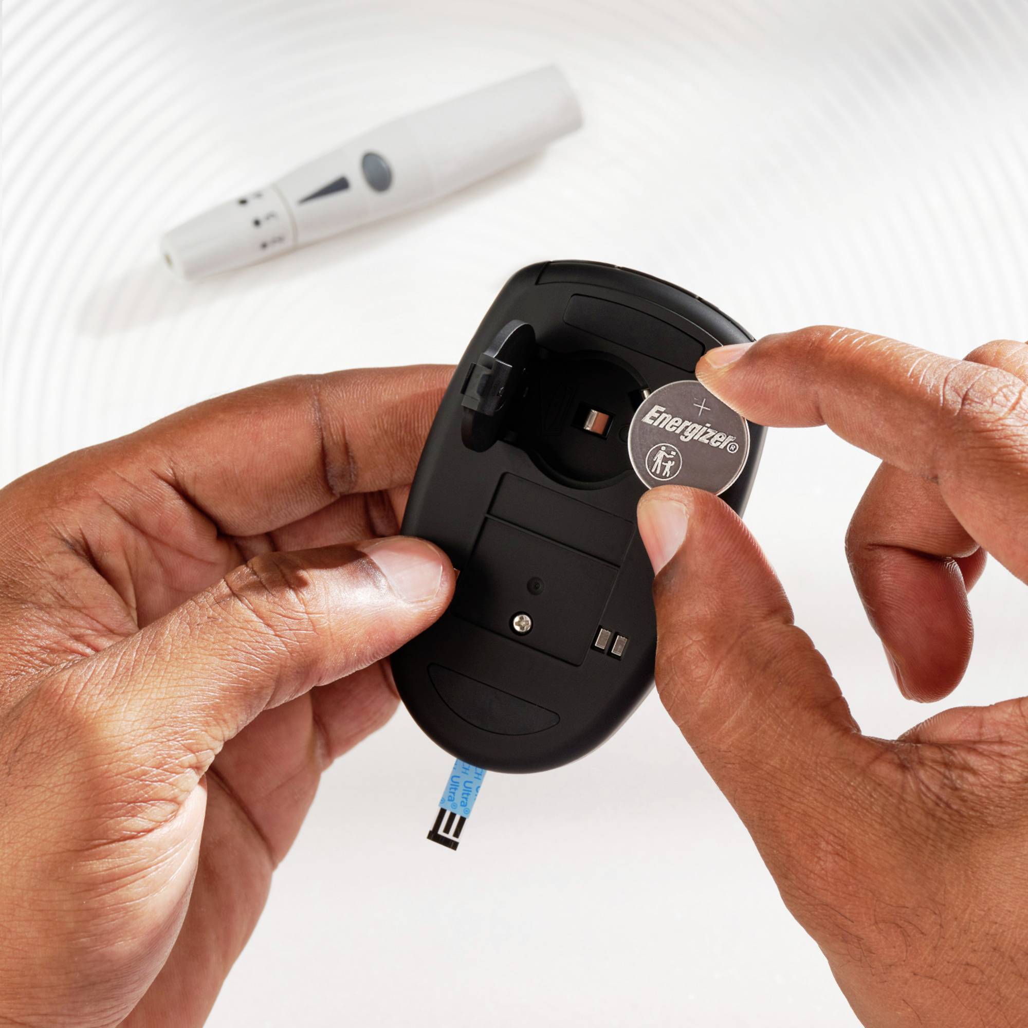 Deux mains tiennent un lecteur de glycémie, une main y insère une pile Energizer. En arrière-plan, on aperçoit un stylo autopiqueur pour diabétiques.