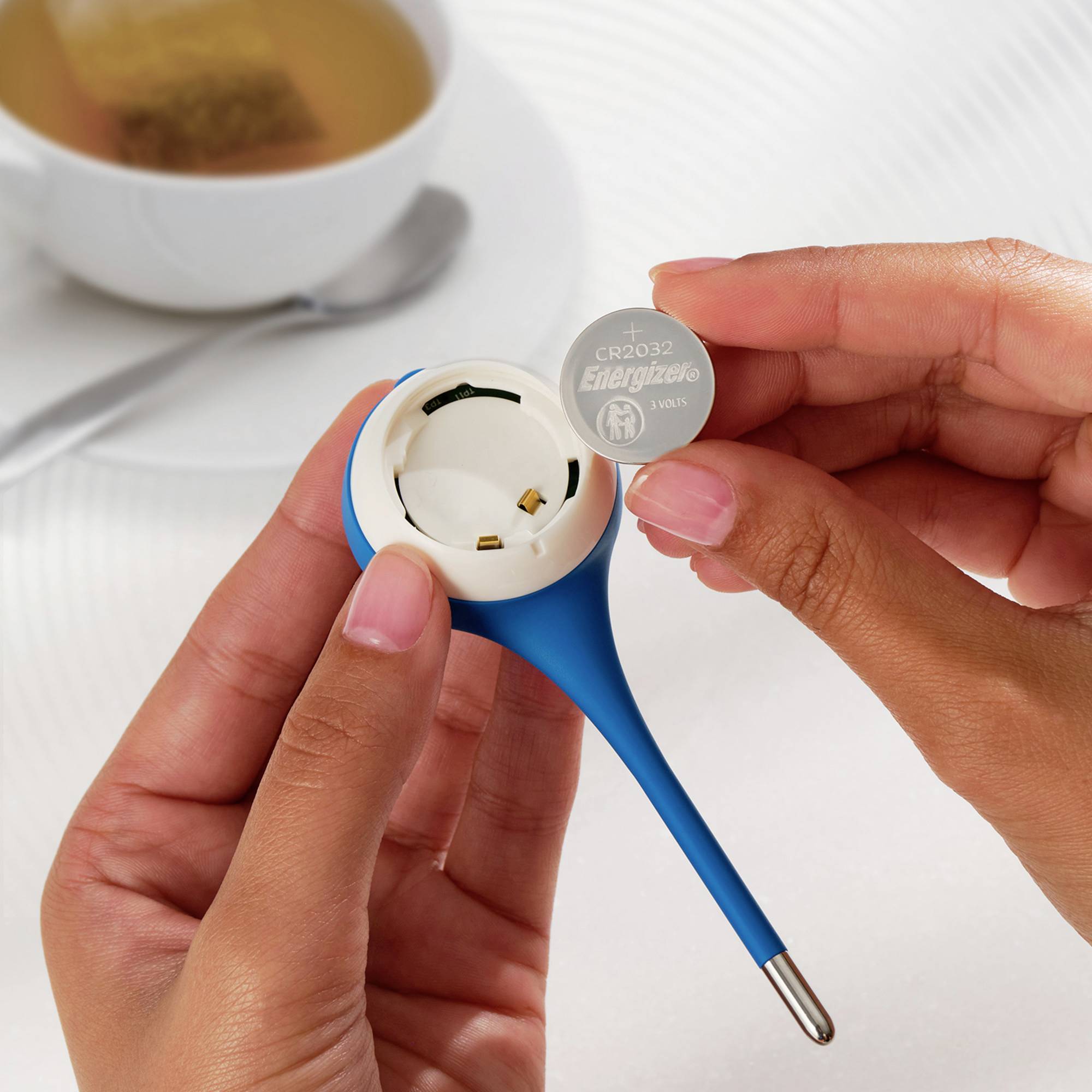 Une personne tient une pile bouton de marque 'Energizer CR2032' à côté d'un boîtier d'appareil bleu sur une table.