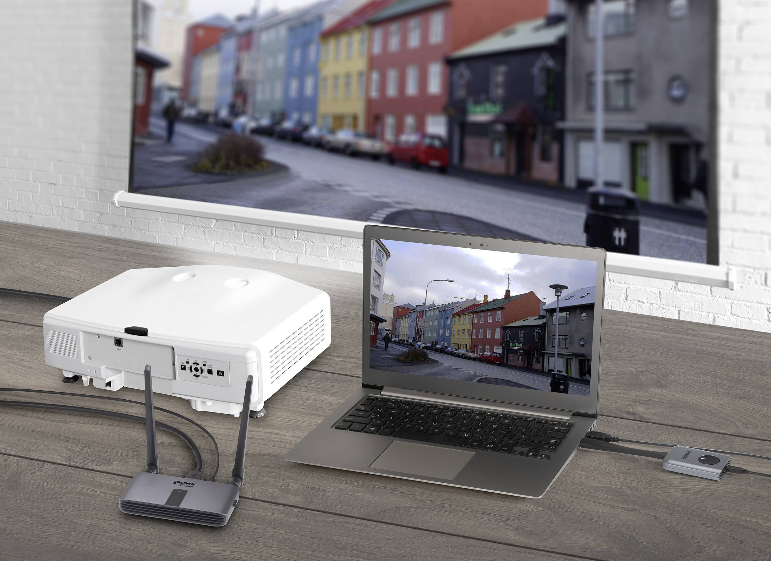 Un projecteur affiche l'image d'une rangée de maisons colorées sur un écran. Un ordinateur portable montre la même image. Configuration technique sur une table en bois.