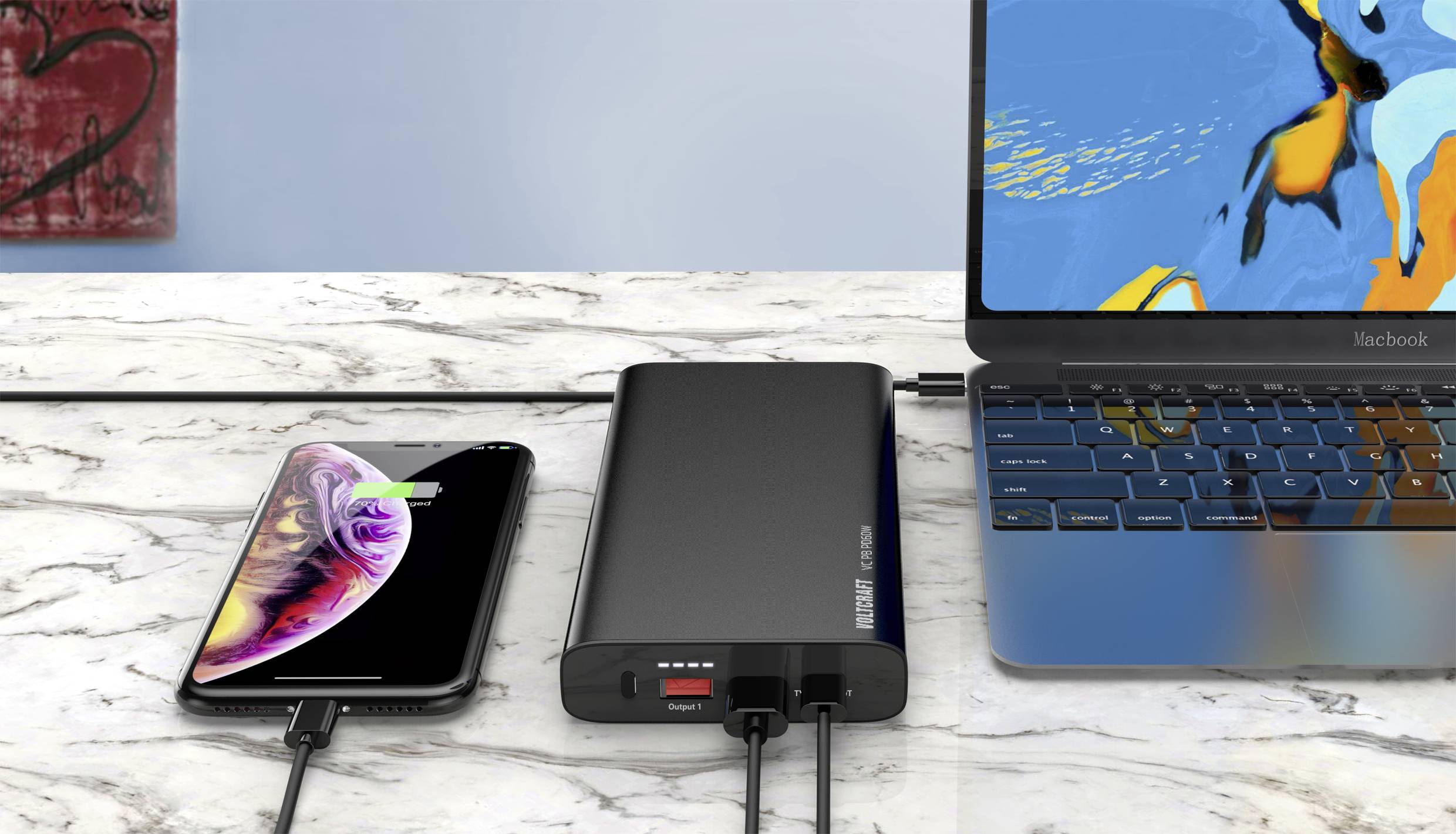 Un smartphone et un ordinateur portable se chargent simultanément avec une batterie externe sur une table en marbre. L'écran du smartphone affiche l'état de la charge.