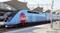 Un train à grande vitesse bleu portant l'inscription 'Ouigo' est stationné en gare. Un toit de gare et un autre train sont visibles sur la gauche.
