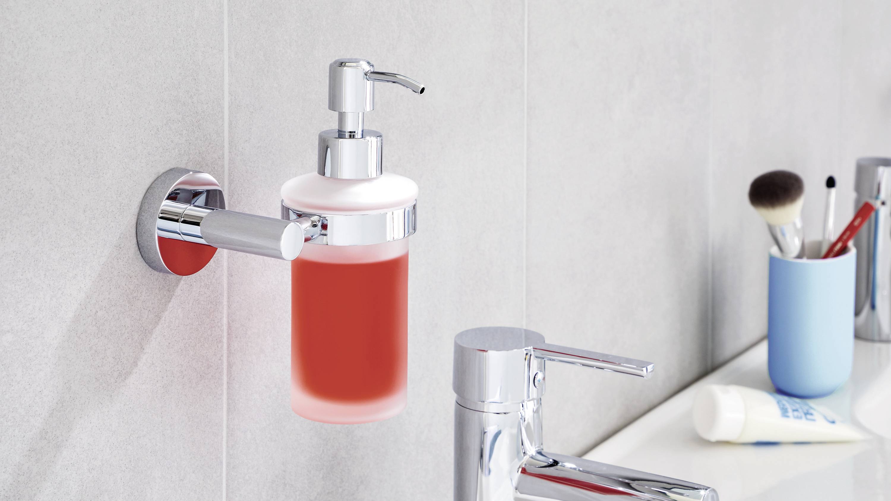 Distributeur de savon liquide rouge fixé au mur. À côté, un robinet et un gobelet contenant des brosses à dents posé sur le lavabo.