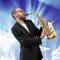 Un homme en costume joue du saxophone sur une scène avec des lumières colorées en arrière-plan, créant une ambiance joyeuse et dynamique.