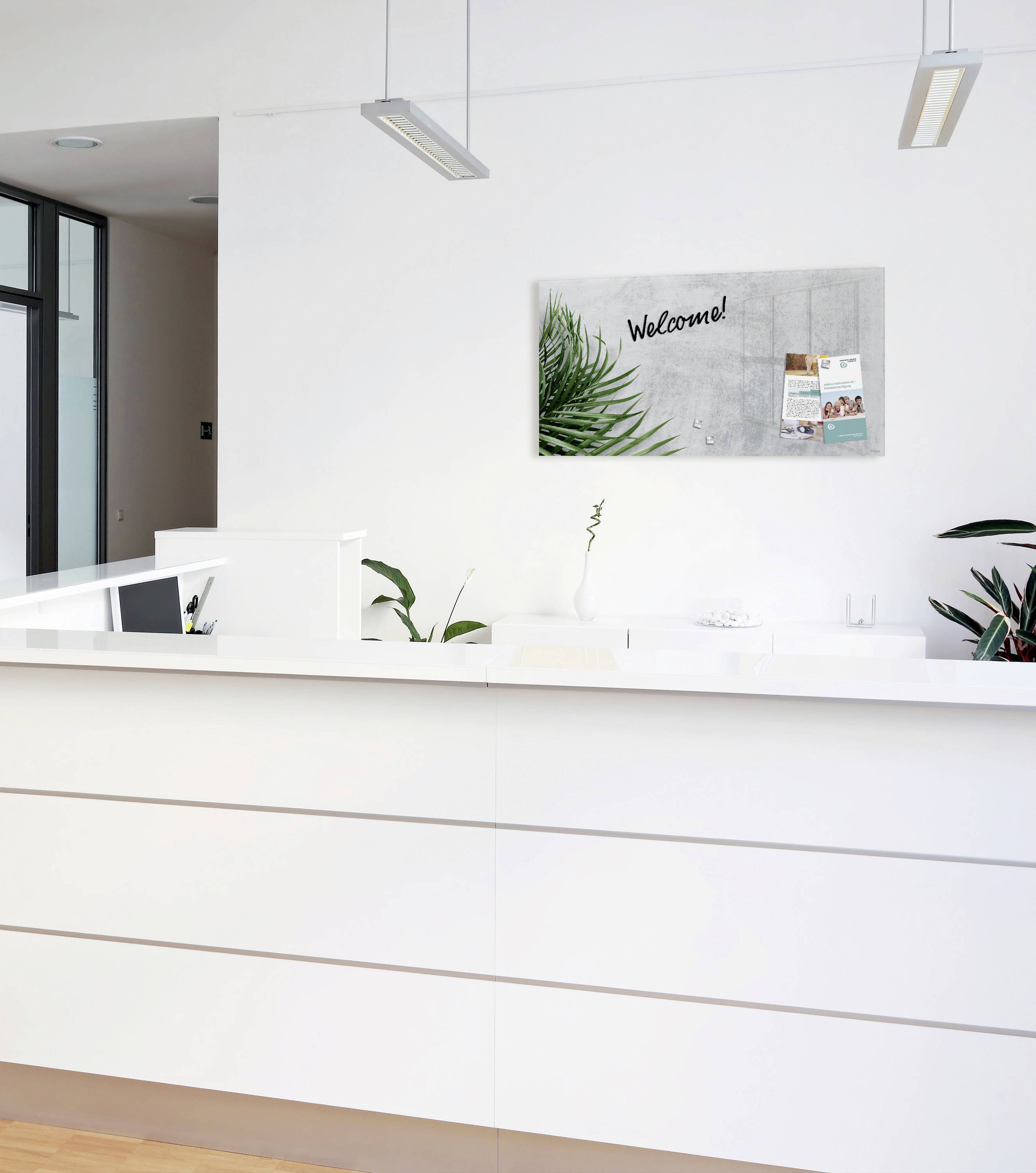 Zone d'accueil avec un comptoir blanc moderne, un tableau 'Welcome!' au-dessus, des plantes et des brochures sur le mur.