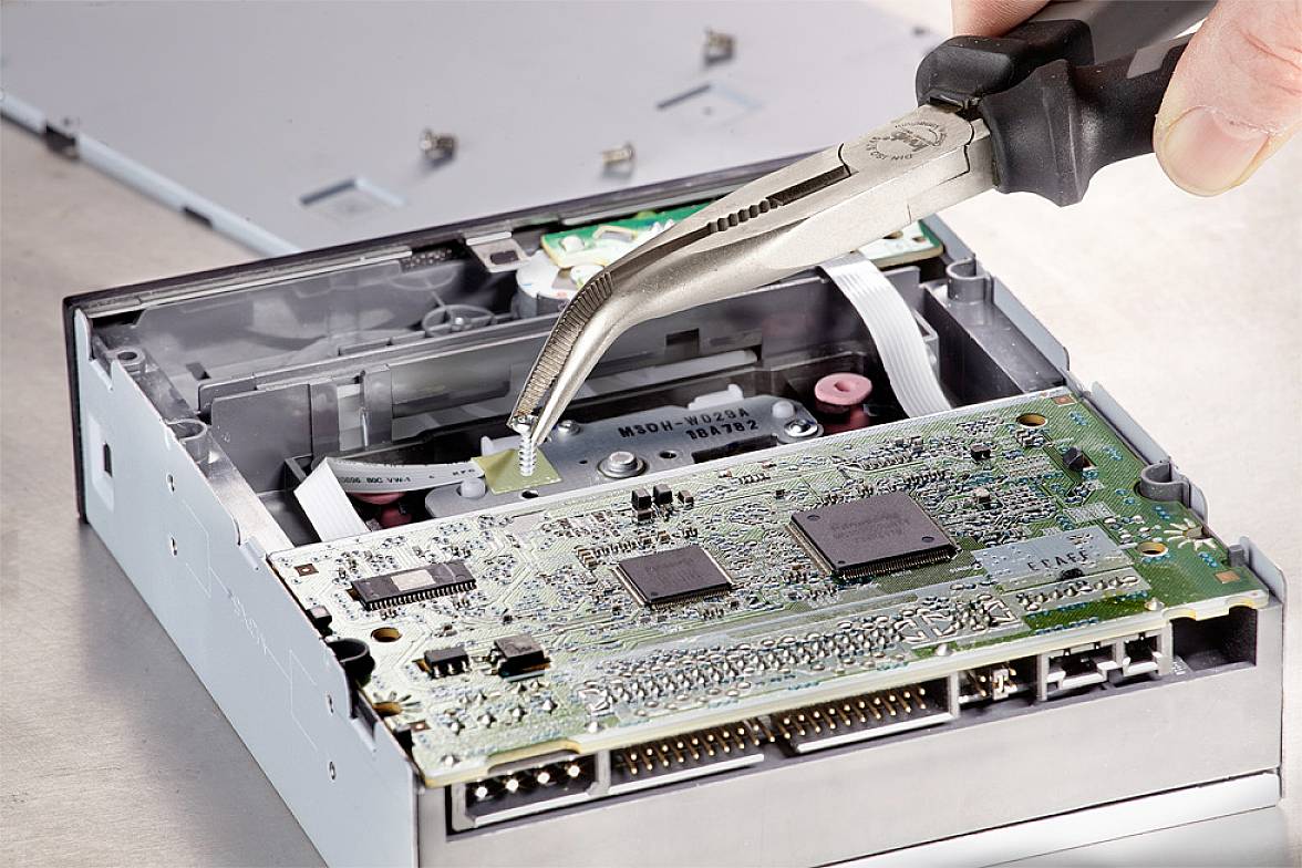 Une main tient une pince brucelles et travaille sur un disque dur d'ordinateur ouvert. La carte mère et les composants électroniques sont visibles.