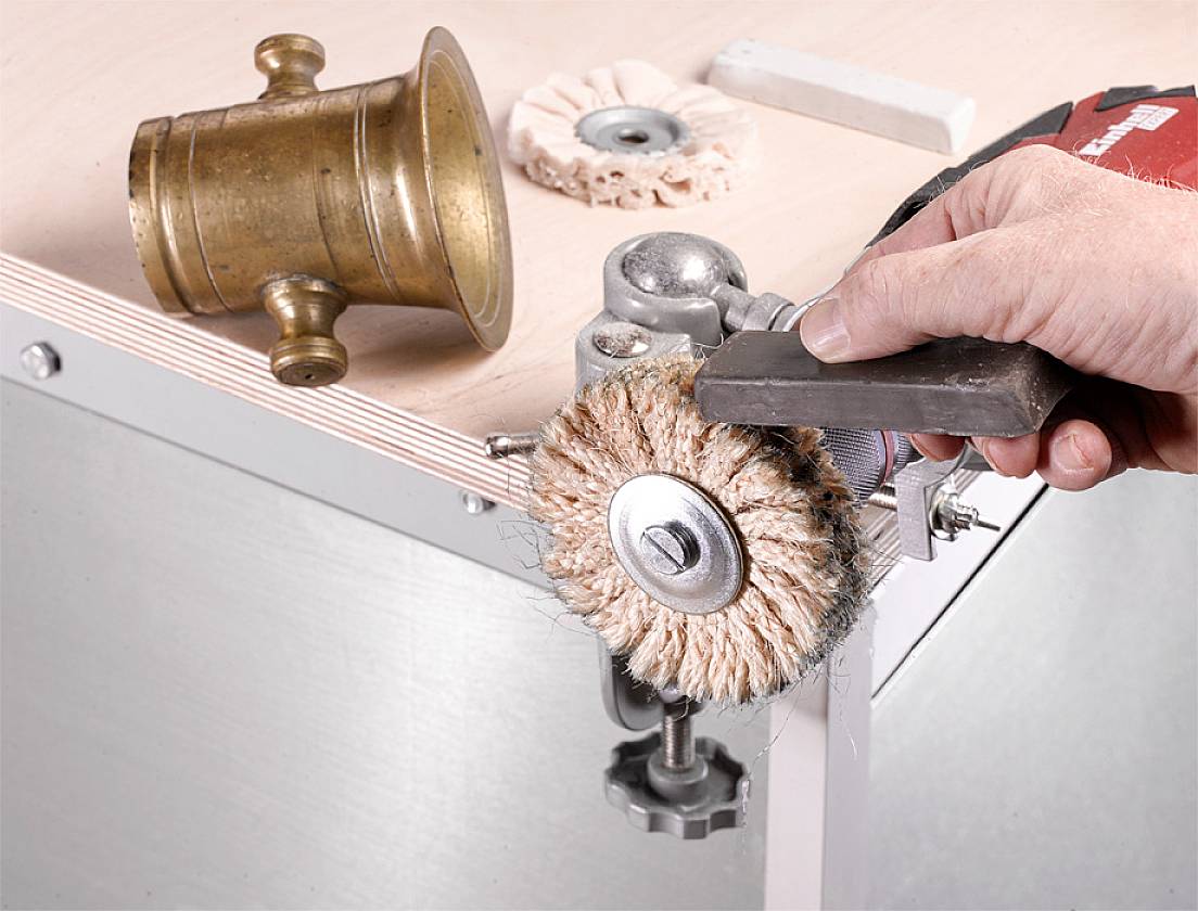 Une main tient un outil avec une brosse métallique, polissant le bord d'une table. Un vieux cor en laiton est posé sur le plateau de la table.
