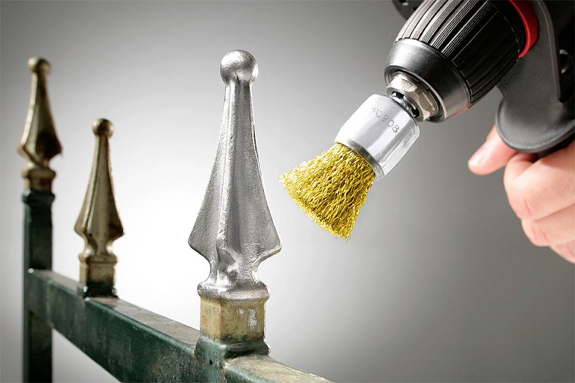Une personne enlève de la rouille d'une rambarde métallique à l'aide d'une brosse métallique fixée à une perceuse, afin de la nettoyer et de la remettre à neuf.