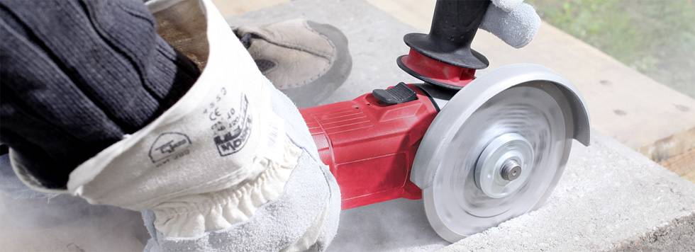 Une personne coupe du béton à l'aide d'une meuleuse d'angle. Elle porte des gants de sécurité, de la poussière visible indique l'activité en cours.