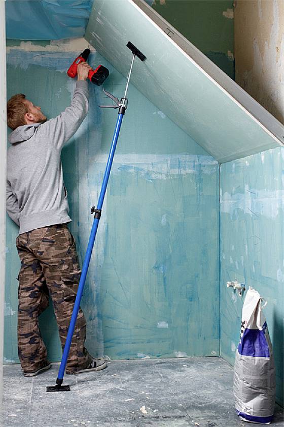 Un homme en tenue de travail installe une plaque de couverture sur un mur de toit incliné dans une pièce aux murs peints en bleu.