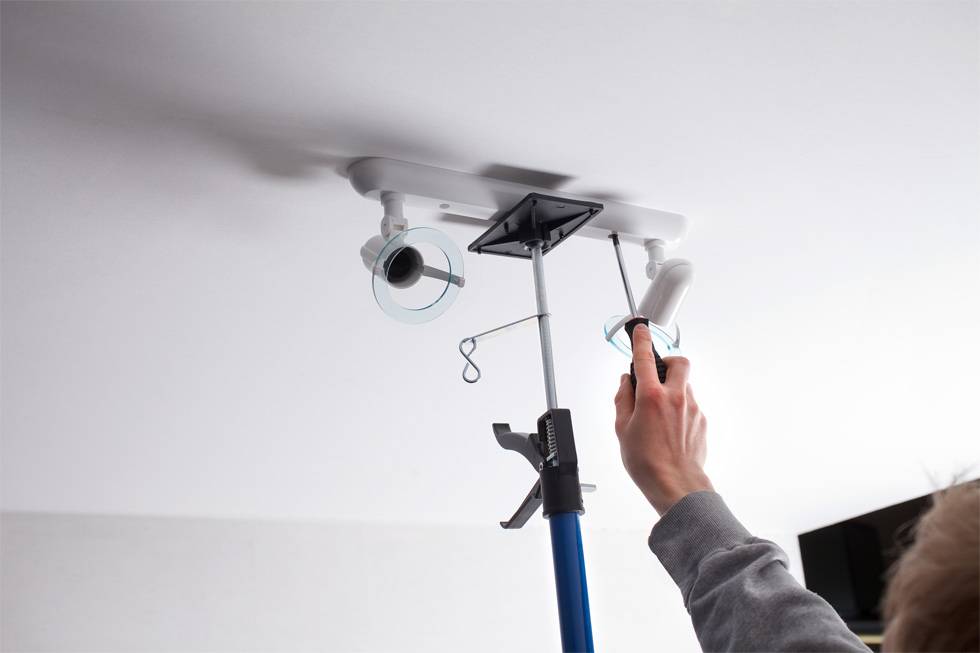 Une personne installe un luminaire plafonnier à deux spots orientables sur un plafond à l'aide d'un outil.