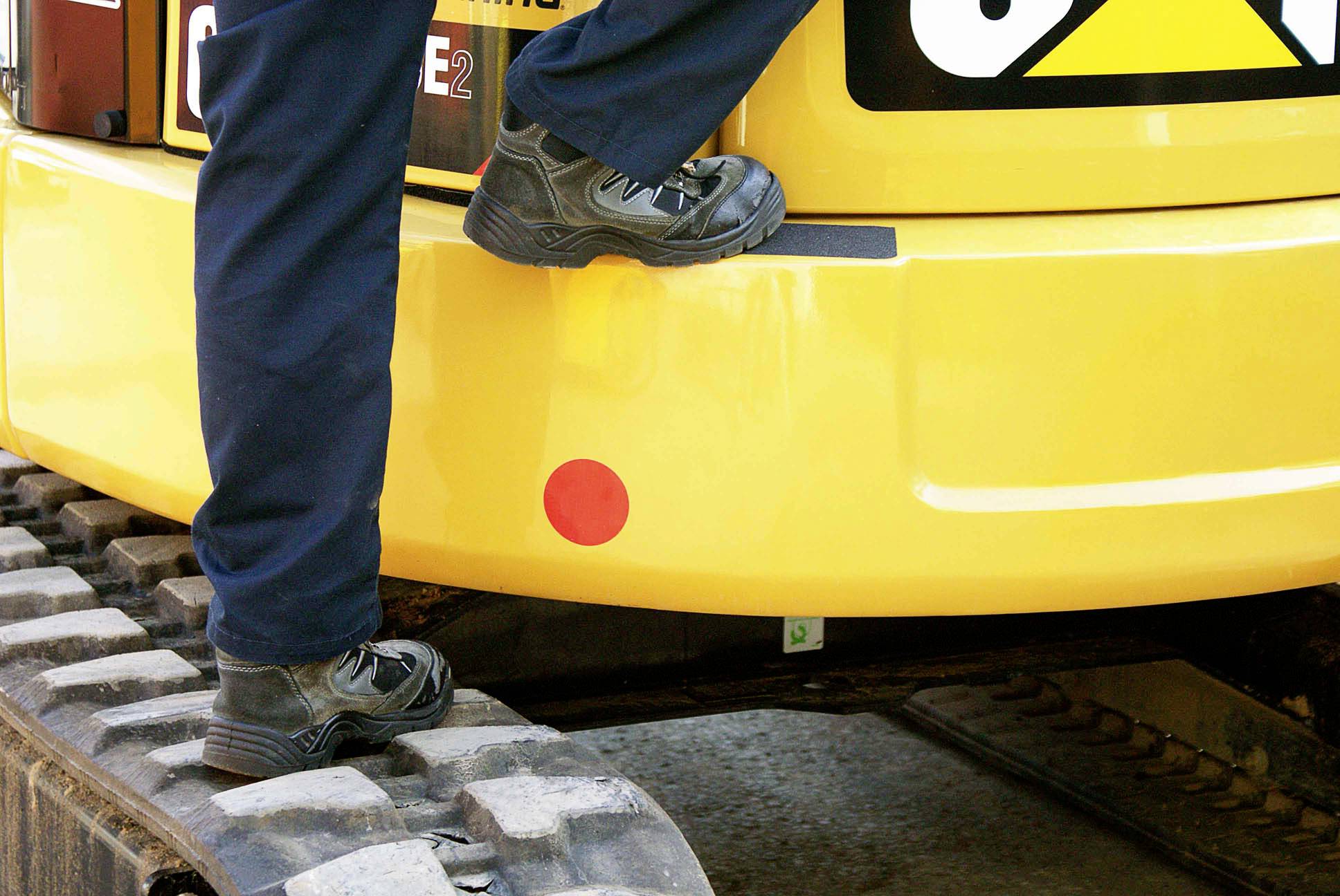 Une personne monte dans un pelleteuse jaune, seules les jambes sont visibles sur les chenilles.
