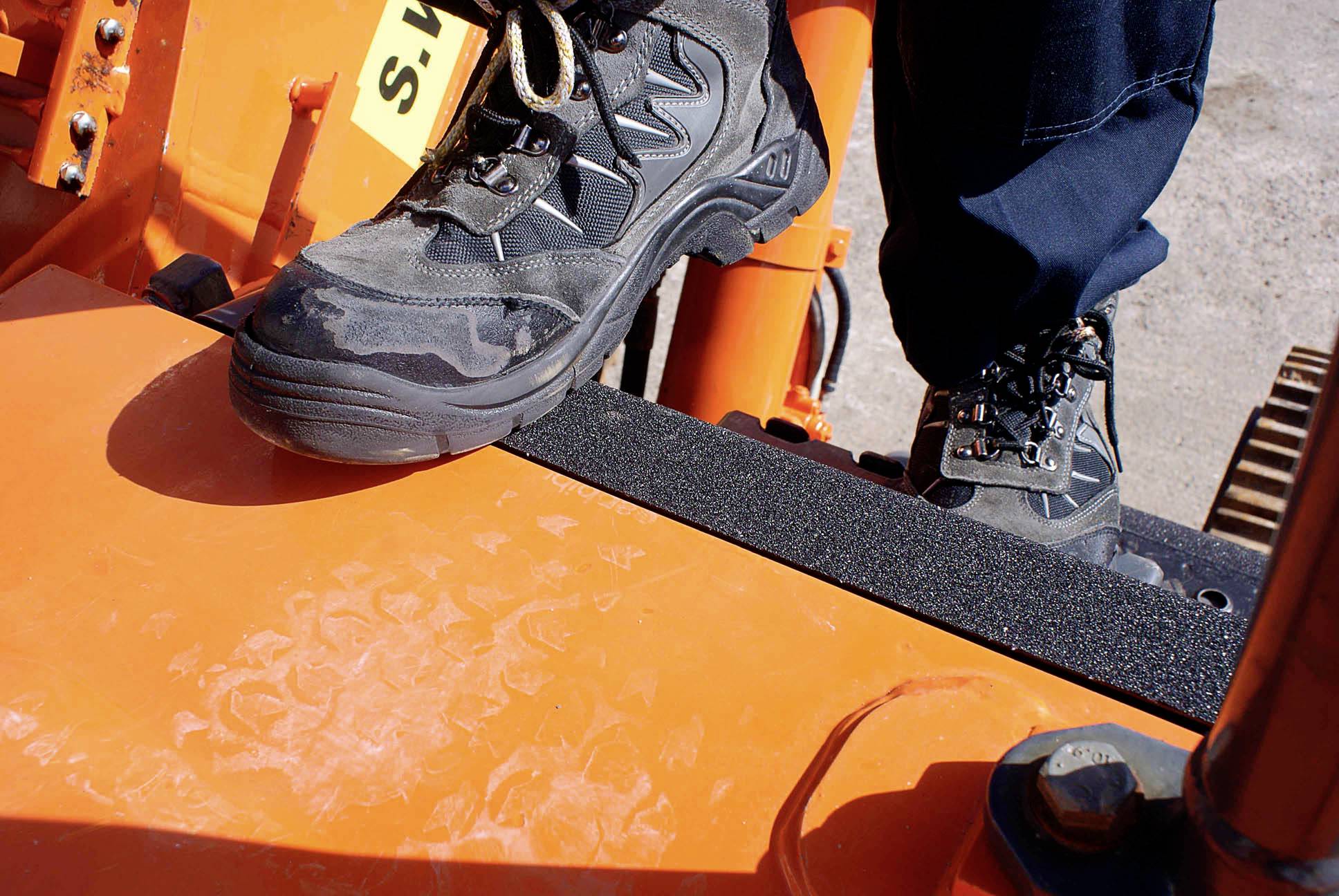 Un ouvrier se tient sur une plateforme orange d'un engin de chantier lourd, chaussé de solides chaussures de randonnée.