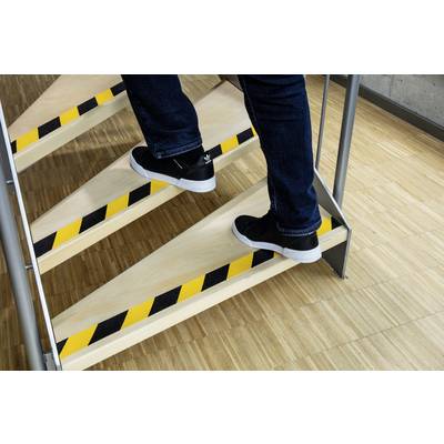 'Gros plan de quelqu'un montant un escalier avec des marquages d'avertissement jaune et noir sur les marches, portant un jean et des baskets noires.'