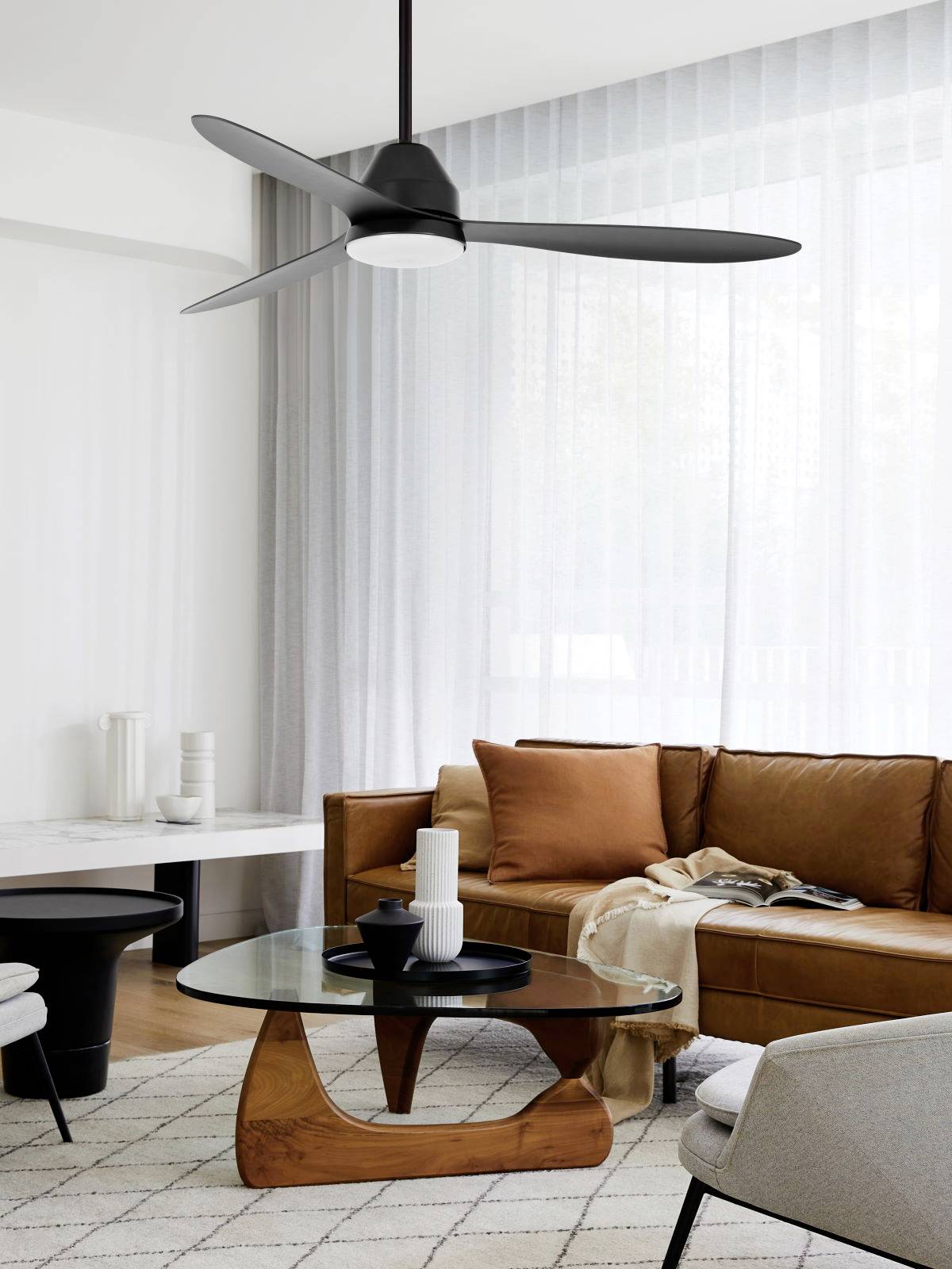 Salon discret avec un canapé en cuir, une table en bois et un ventilateur de plafond moderne. Des rideaux lumineux et un tapis créent une ambiance chaleureuse.