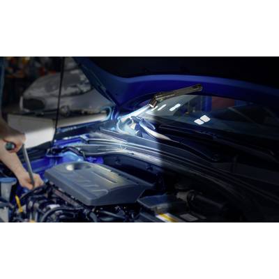 Un mécanicien travaille sous le capot ouvert d'une voiture, éclairé par une lampe. L'accent est mis sur les détails techniques.