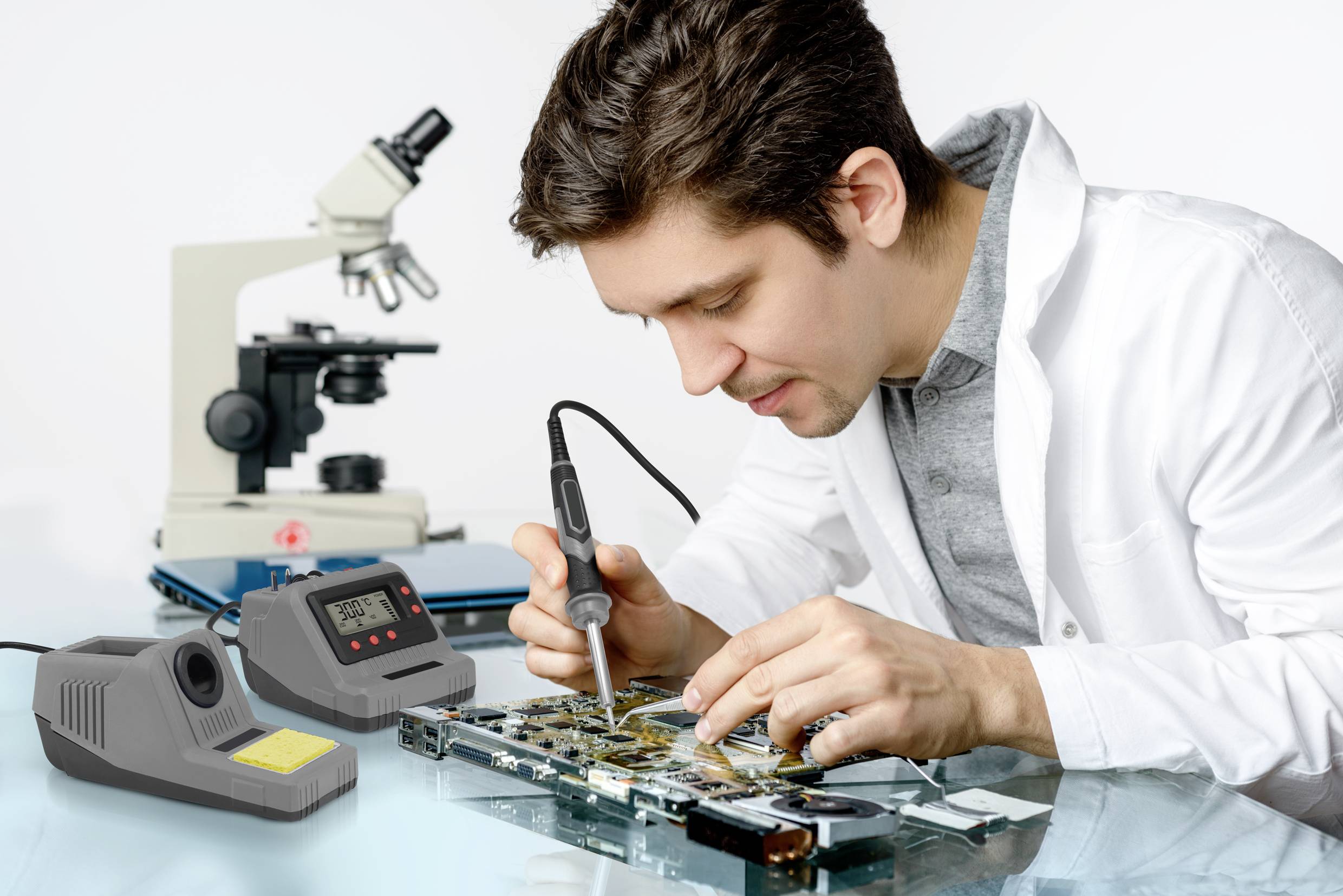 Un homme soude sur une carte de circuit imprimé, un microscope est posé sur une table à côté de lui.