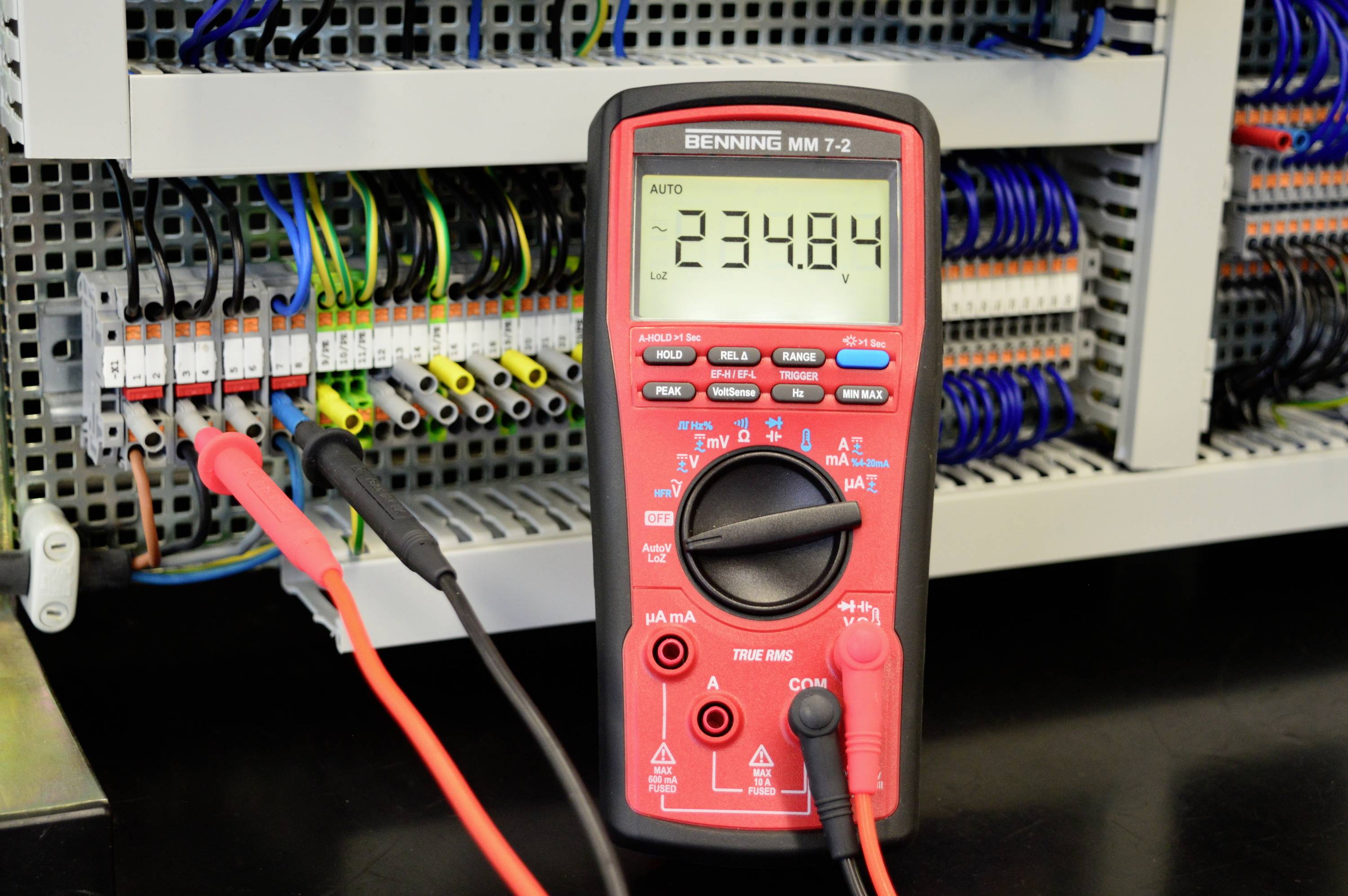 Un multimètre numérique mesure une tension de 234,84 V sur un tableau de distribution ouvert comportant plusieurs câbles et connexions de différentes couleurs.