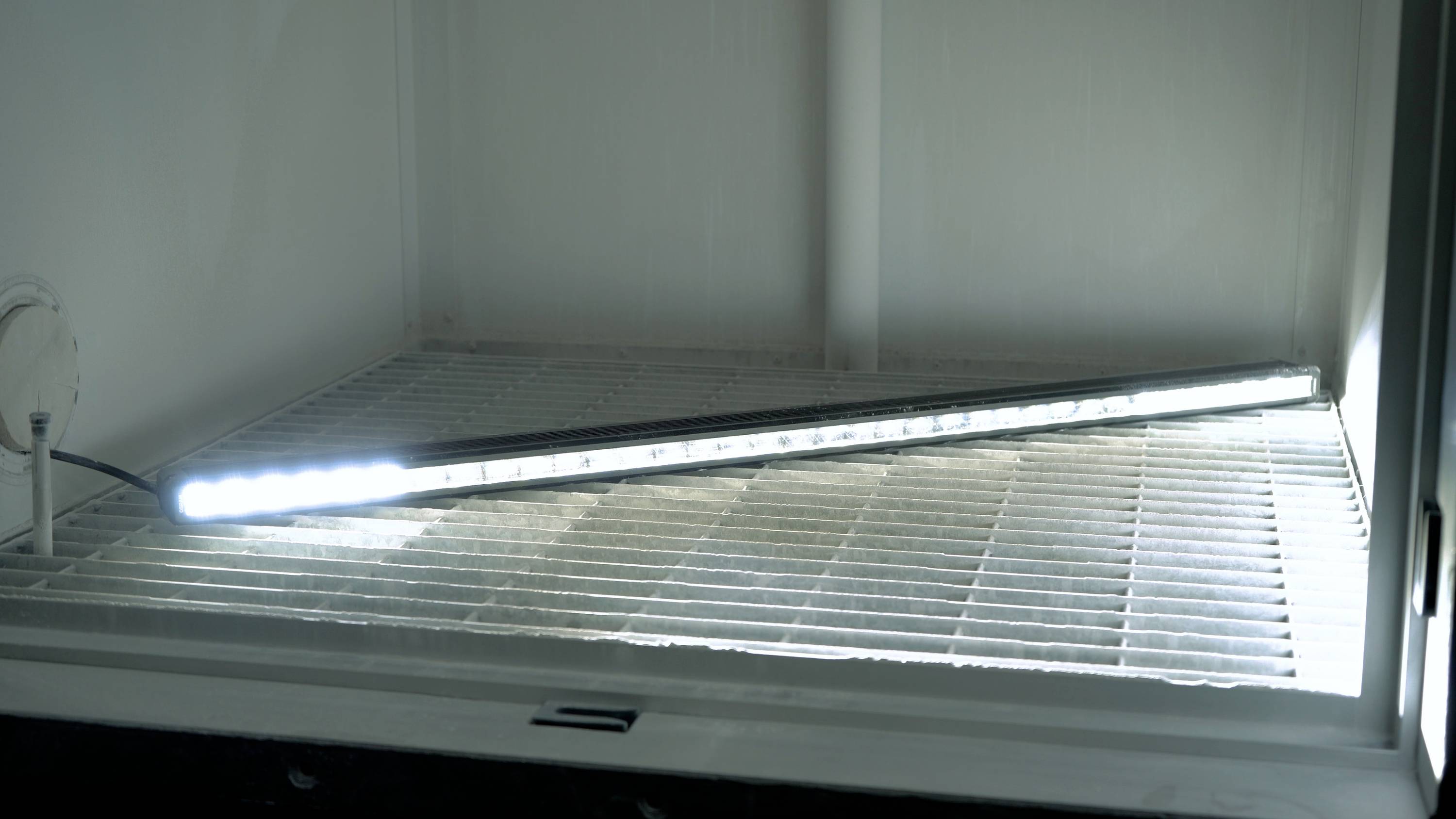 Une longue lampe LED lumineuse est posée en diagonale sur une grille dans une pièce vide et éclairée.
