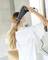 Une femme aux longs cheveux blonds sèche ses cheveux dans une salle de bain. Elle porte un haut blanc. Des fleurs sont floues en arrière-plan.