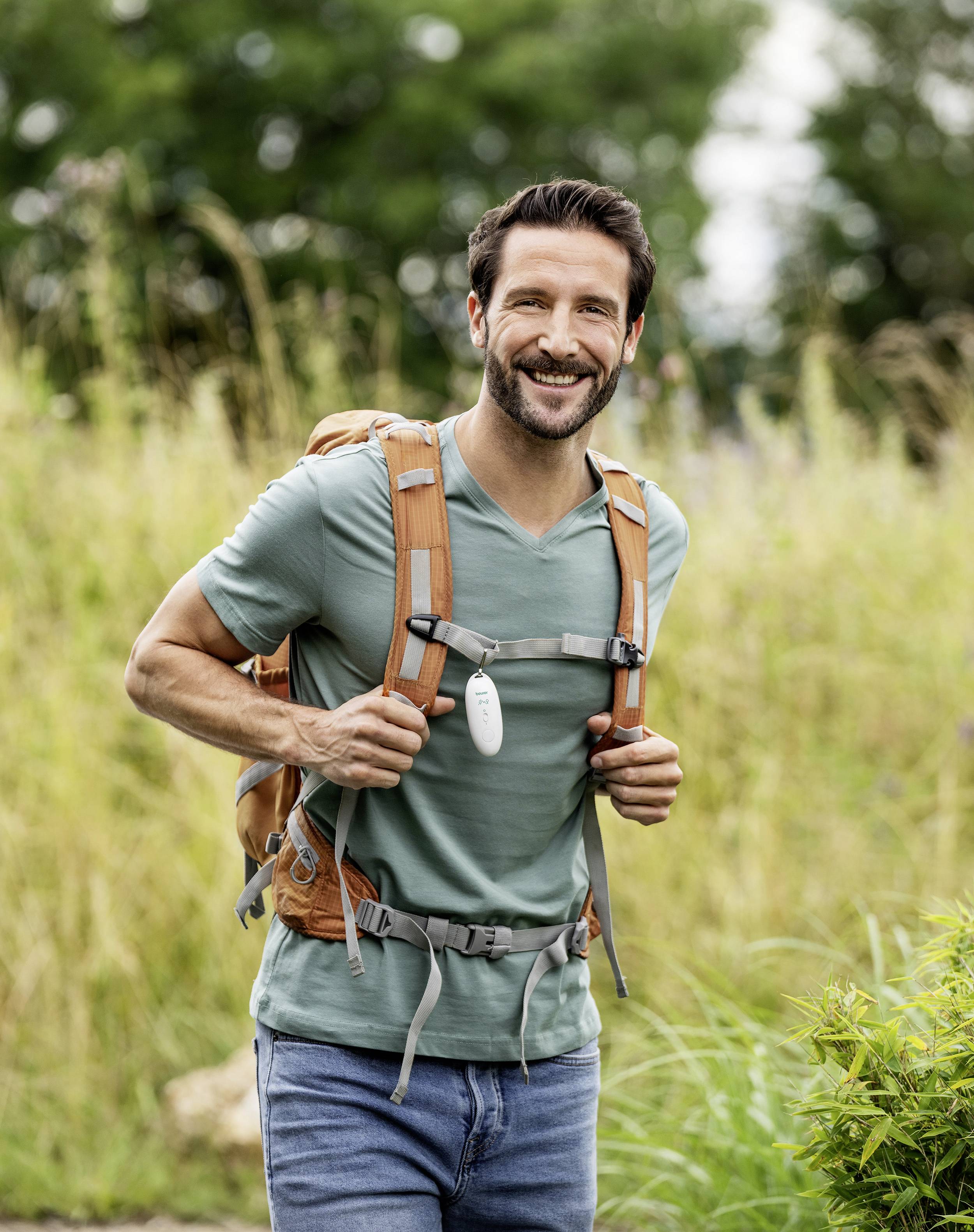 Un homme aux cheveux bruns marche joyeusement sur un chemin verdoyant. Il porte un sac à dos marron et un t-shirt vert.