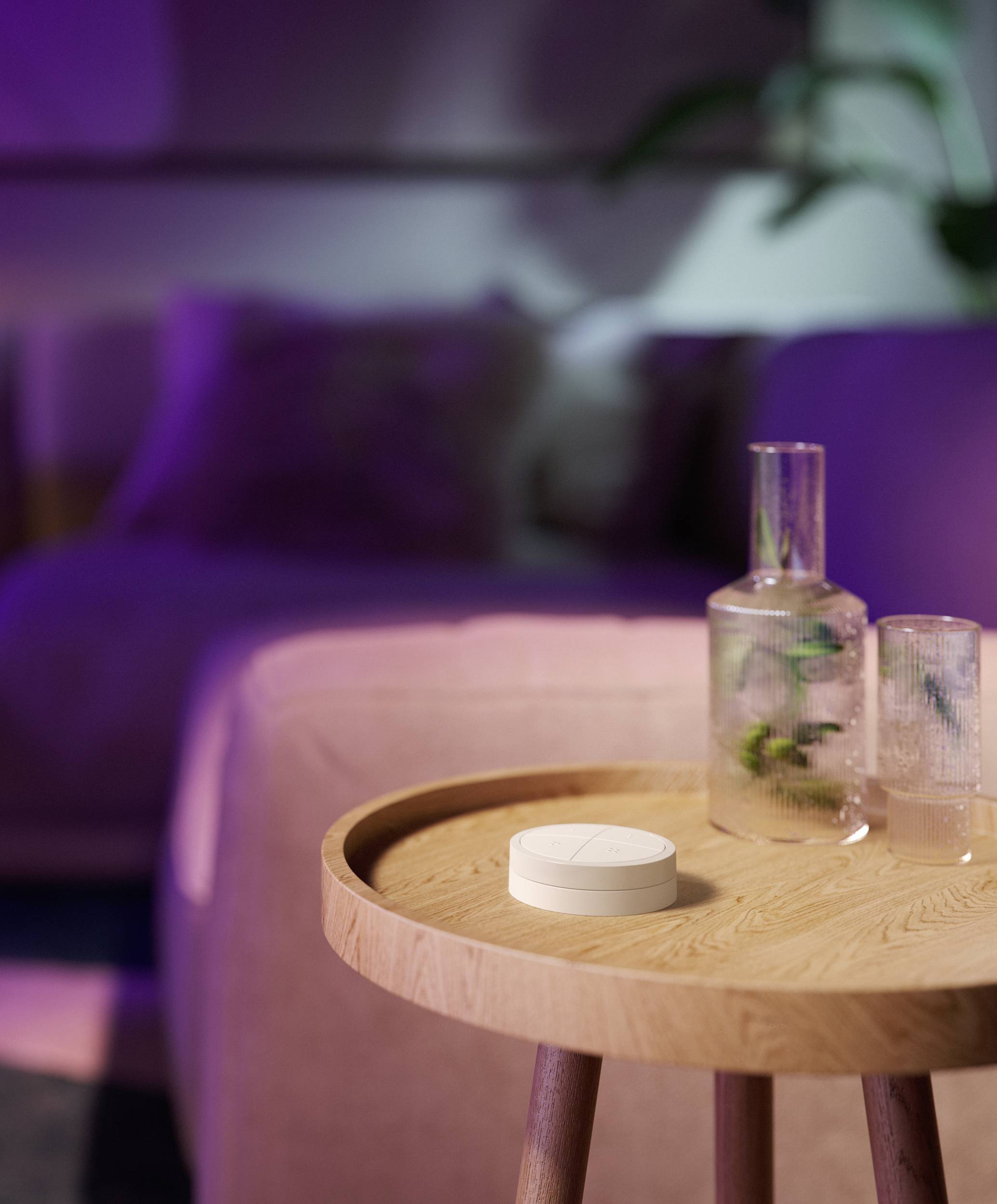 Une table ronde en bois avec un petit appareil blanc posé dessus, à côté de deux verres. En arrière-plan, une ambiance de salon chaleureuse et floue.