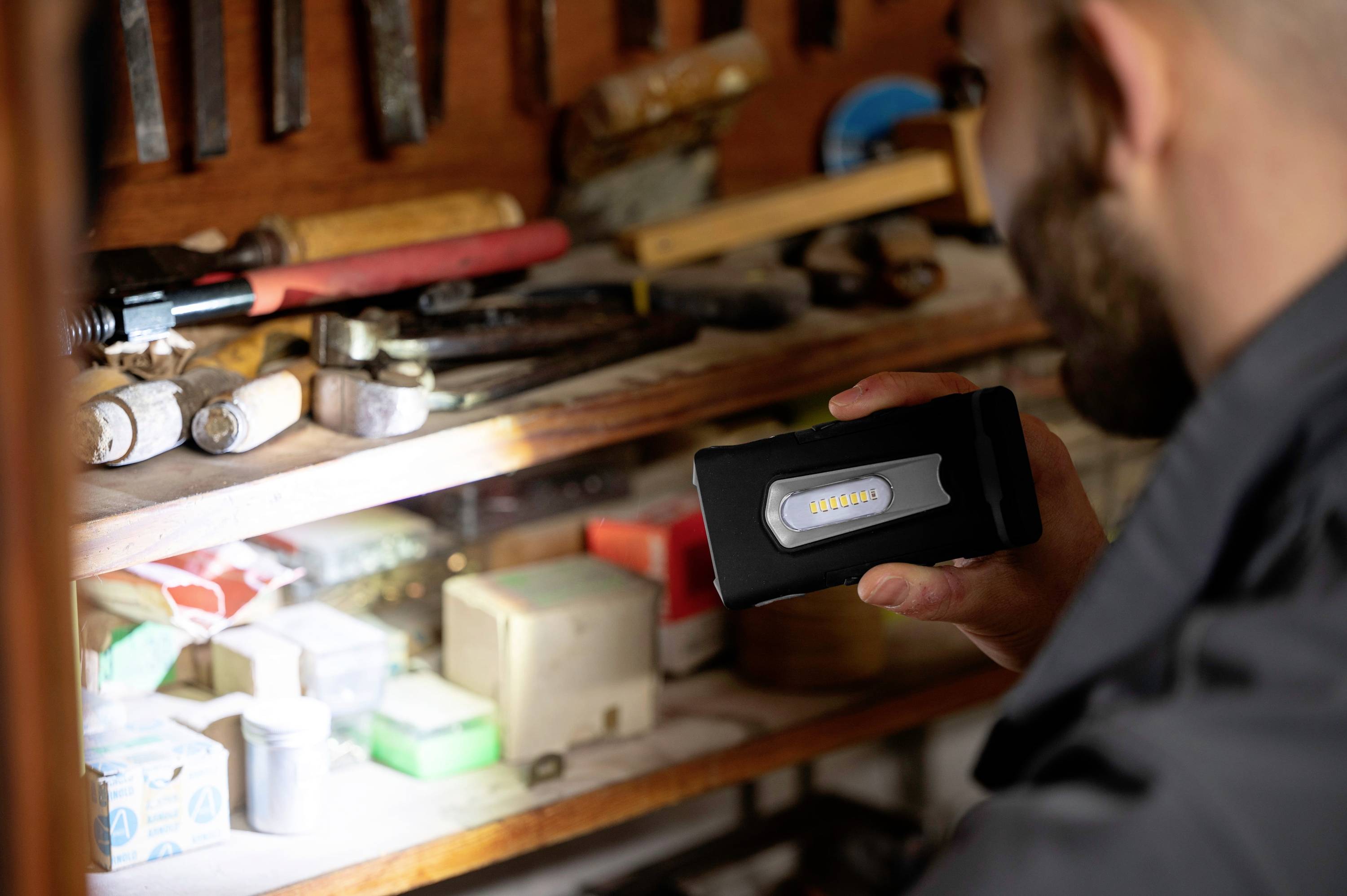 Un homme tient une lampe de poche et éclaire une étagère remplie d'outils. Sur l'étagère se trouvent différents outils et cartons.