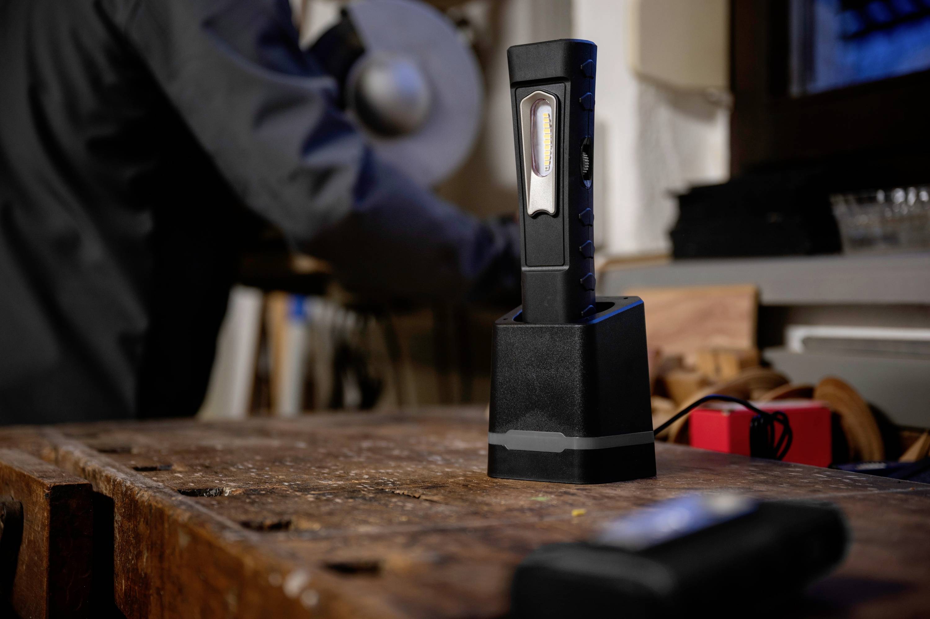 Une lampe de travail noire est posée sur un établi. À l'arrière-plan, on aperçoit une personne floue travaillant sur un outil.