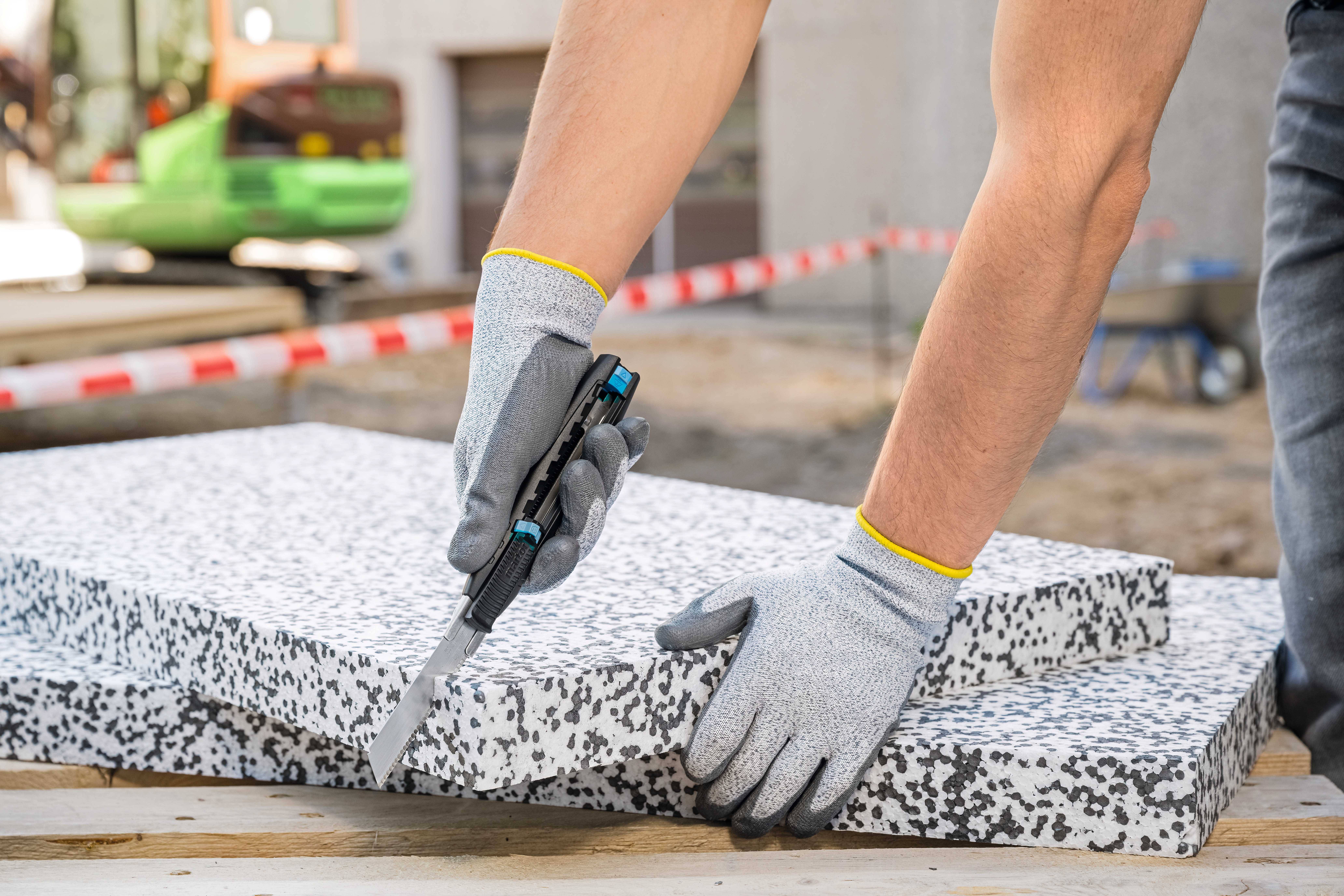 Une personne portant des gants gris coupe une plaque de polystyrène avec un couteau. Un chantier de construction est visible en arrière-plan.