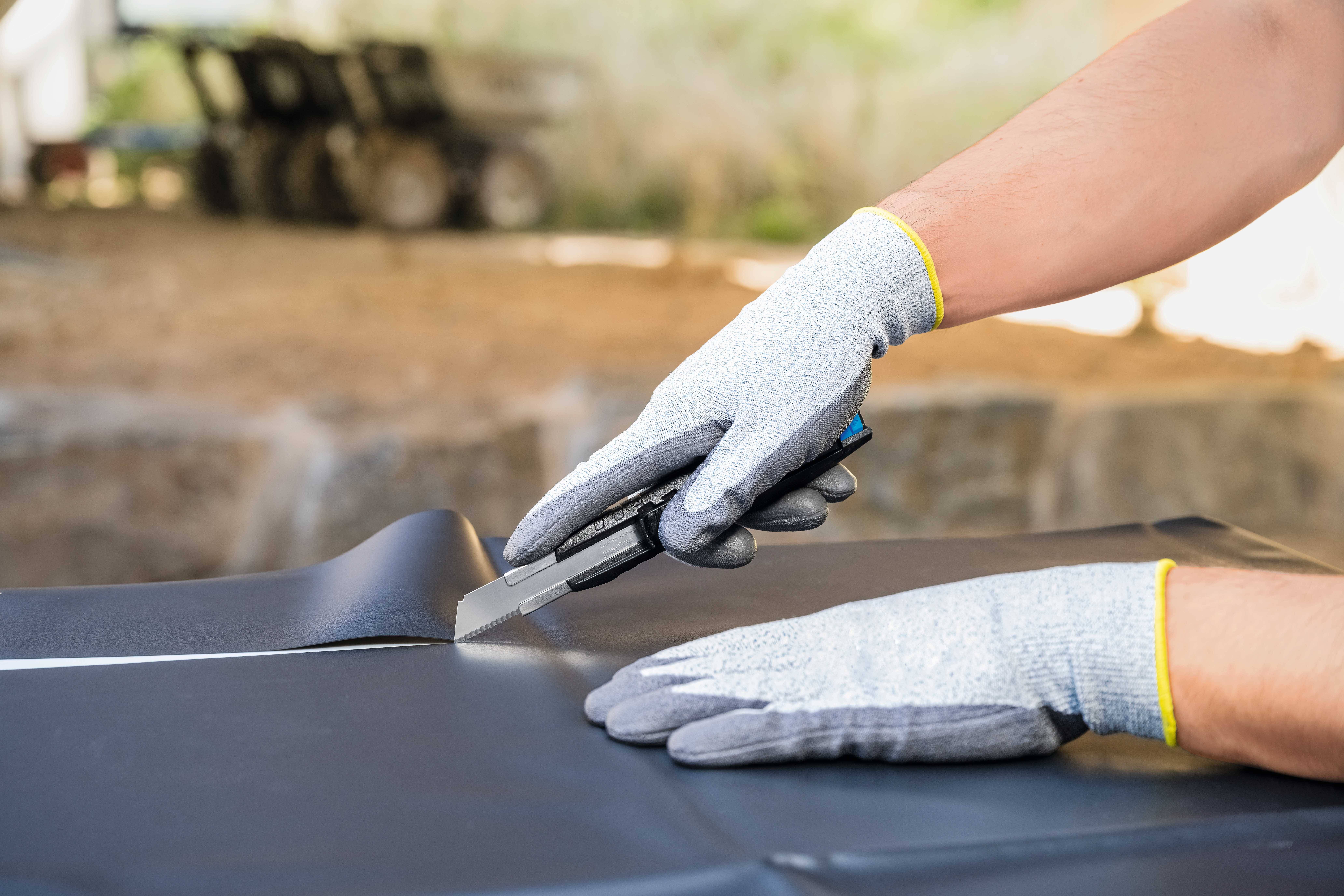 Une personne portant des gants de travail gris découpe du matériau noir avec un cutter. L'arrière-plan est flou.
