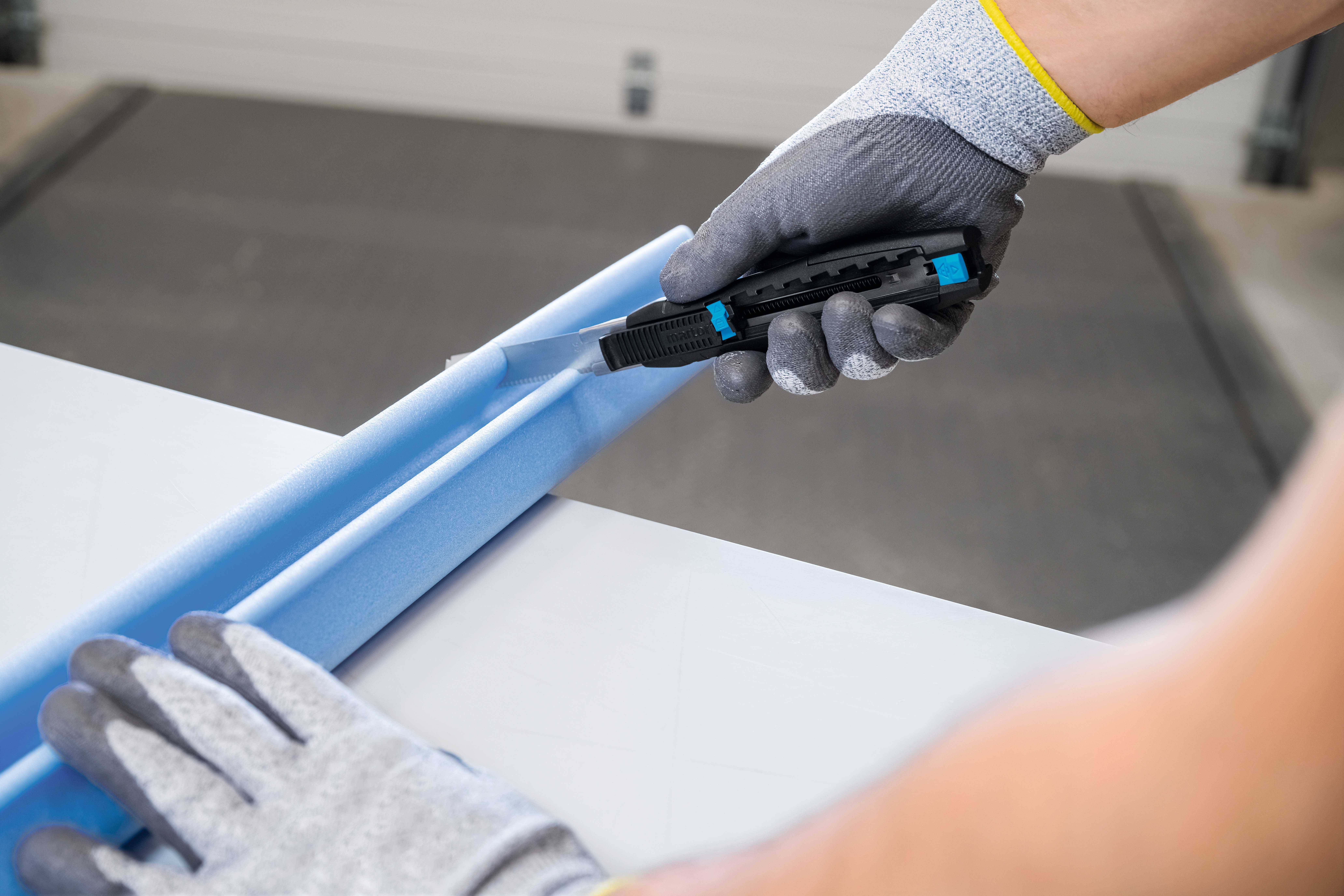 Une personne coupe du mousse bleu avec un cutter. Elle porte des gants gris.